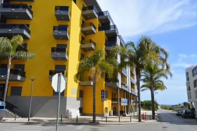 a yellow building with palm trees in front of it at Apartamento frente ao mar com piscina de água salgada in Quarteira